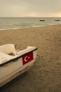 Çatalca’da Yazın Gidilecek Plajlar 15 Çatalca sahilinde kıyıya çekilmiş Türk bayraklı küçük bir tekne, kum plaj ve denizde duran iki balıkçı teknesi.