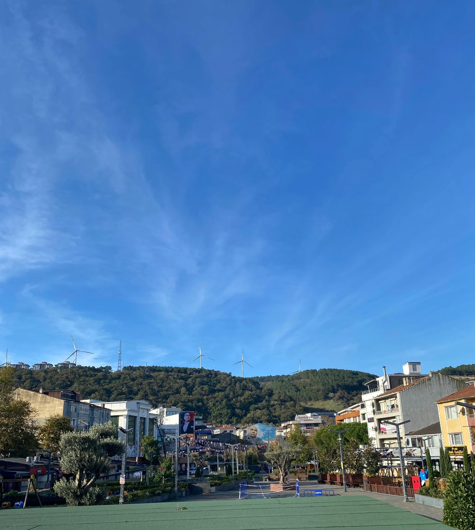 Şehir merkezinde açık bir meydan, arka planda ormanlık tepe ve üzerinde rüzgar türbinleri, altında modern binalar ve açık mavi gökyüzü